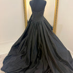 A black ballgown displayed on a black dress form mannequin, showing the back view of the gown with its full train spread across a light-colored floor in front of a white wall with a gold-framed mirror.