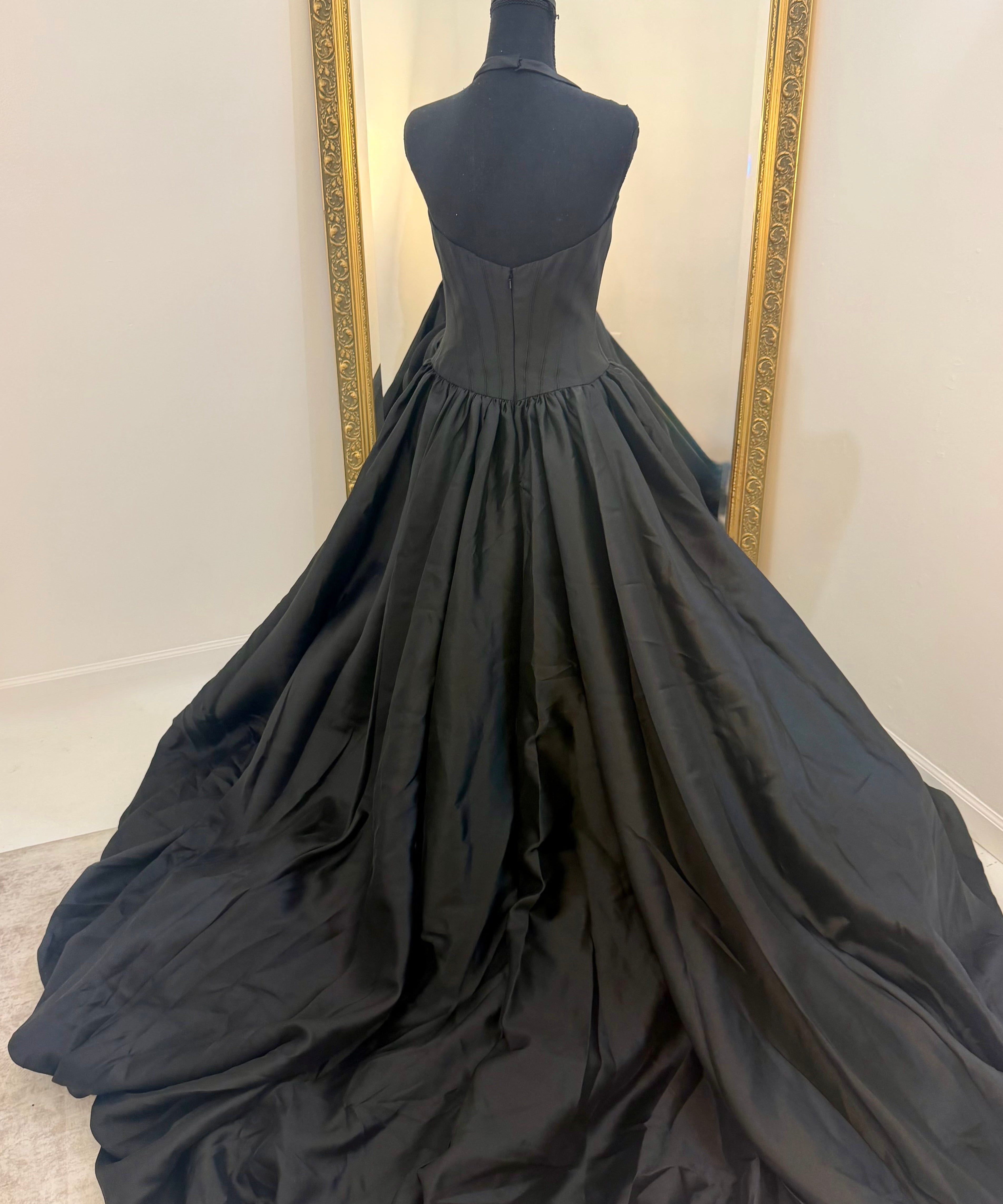 A black ballgown displayed on a black dress form mannequin, showing the back view of the gown with its full train spread across a light-colored floor in front of a white wall with a gold-framed mirror.