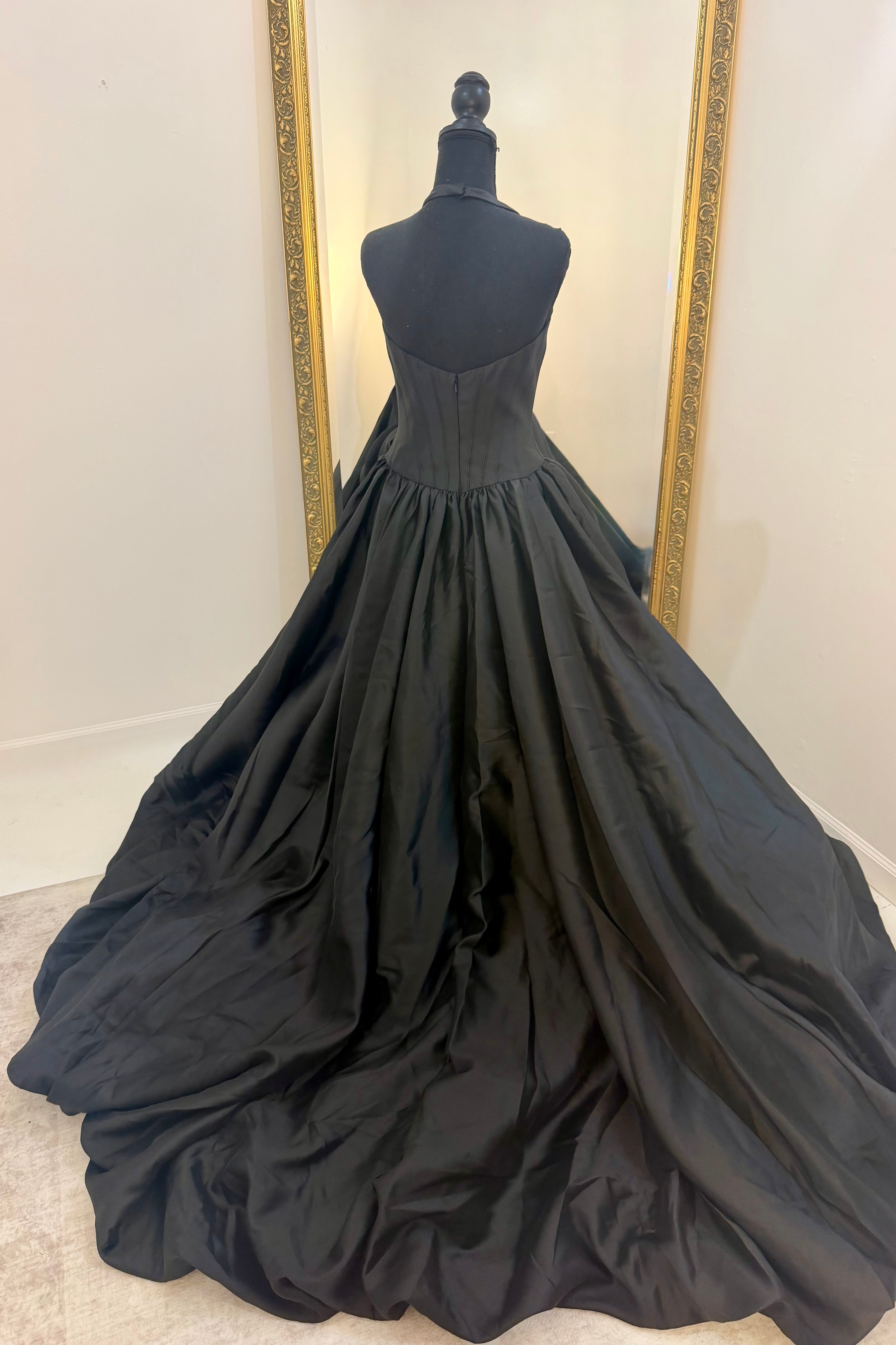 A black ballgown displayed on a black dress form mannequin, showing the back view of the gown with its full train spread across a light-colored floor in front of a white wall with a gold-framed mirror.