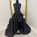 A black formal gown displayed on a black mannequin in front of a large gold-framed mirror. The dress features a sleeveless plunging bodice, a voluminous gathered skirt, and a large sculptural rose detail on the right side of the skirt.
