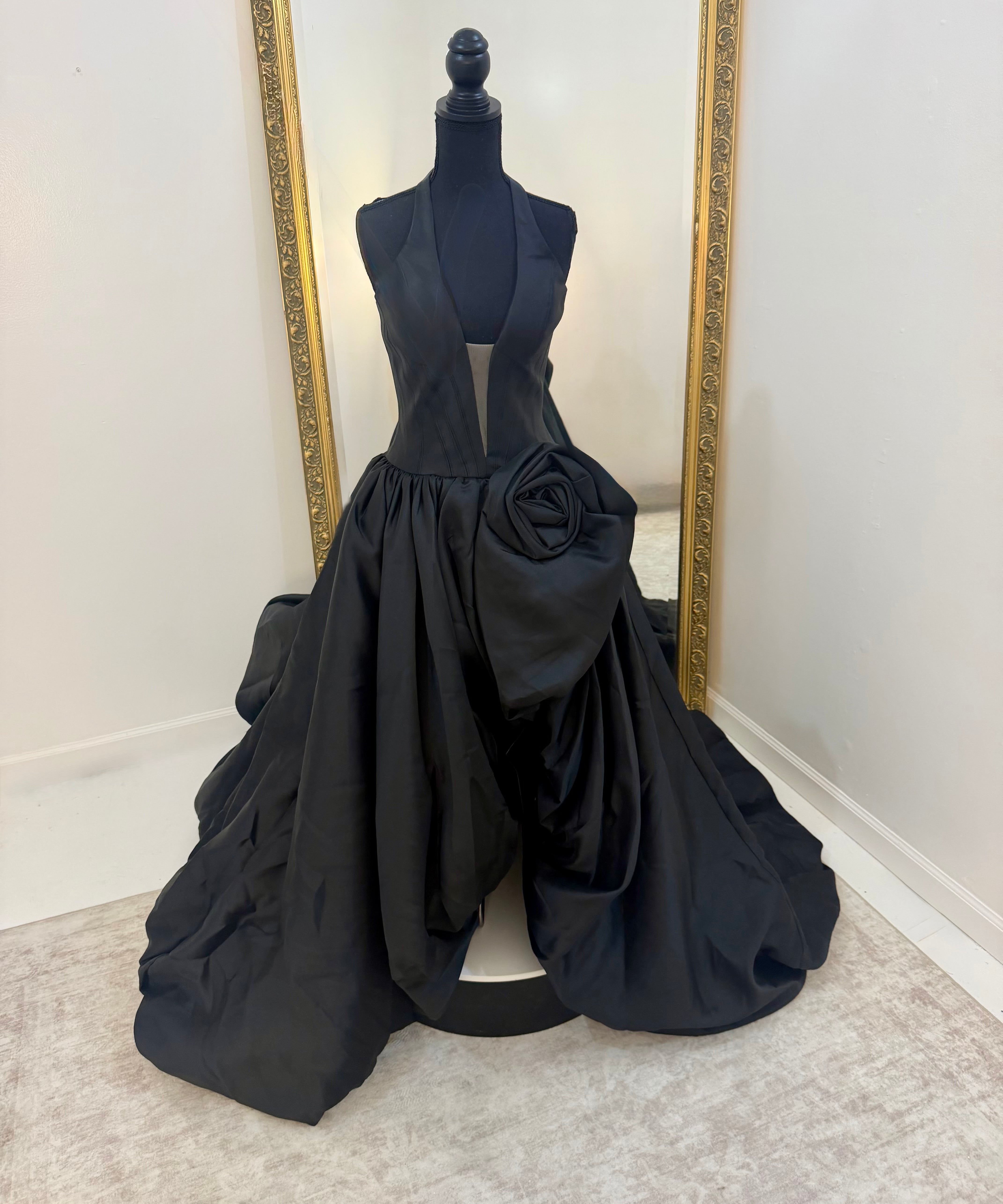 A black formal gown displayed on a black mannequin in front of a large gold-framed mirror. The dress features a sleeveless plunging bodice, a voluminous gathered skirt, and a large sculptural rose detail on the right side of the skirt.