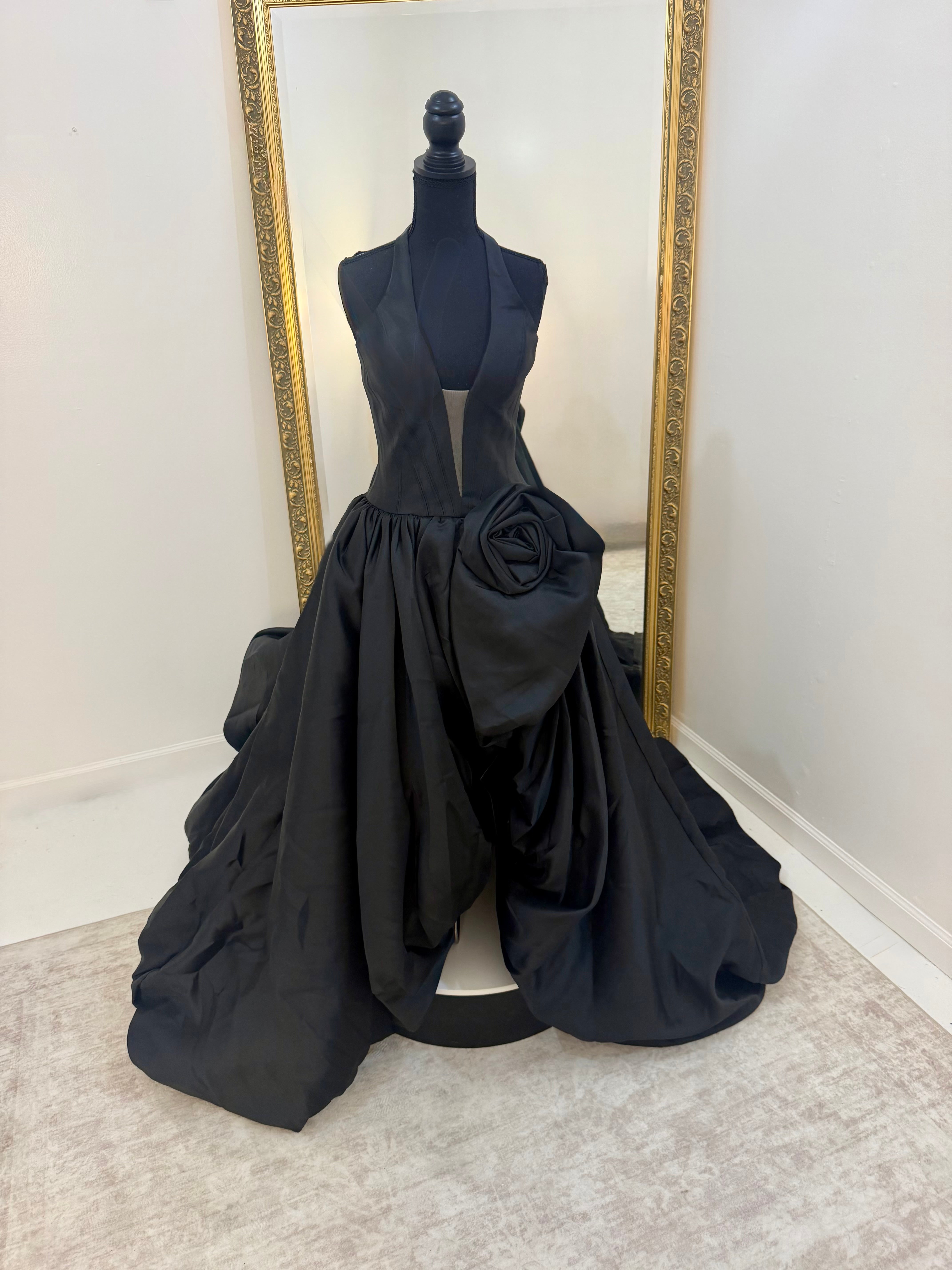 A black formal gown displayed on a black mannequin in front of a large gold-framed mirror. The dress features a sleeveless plunging bodice, a voluminous gathered skirt, and a large sculptural rose detail on the right side of the skirt.