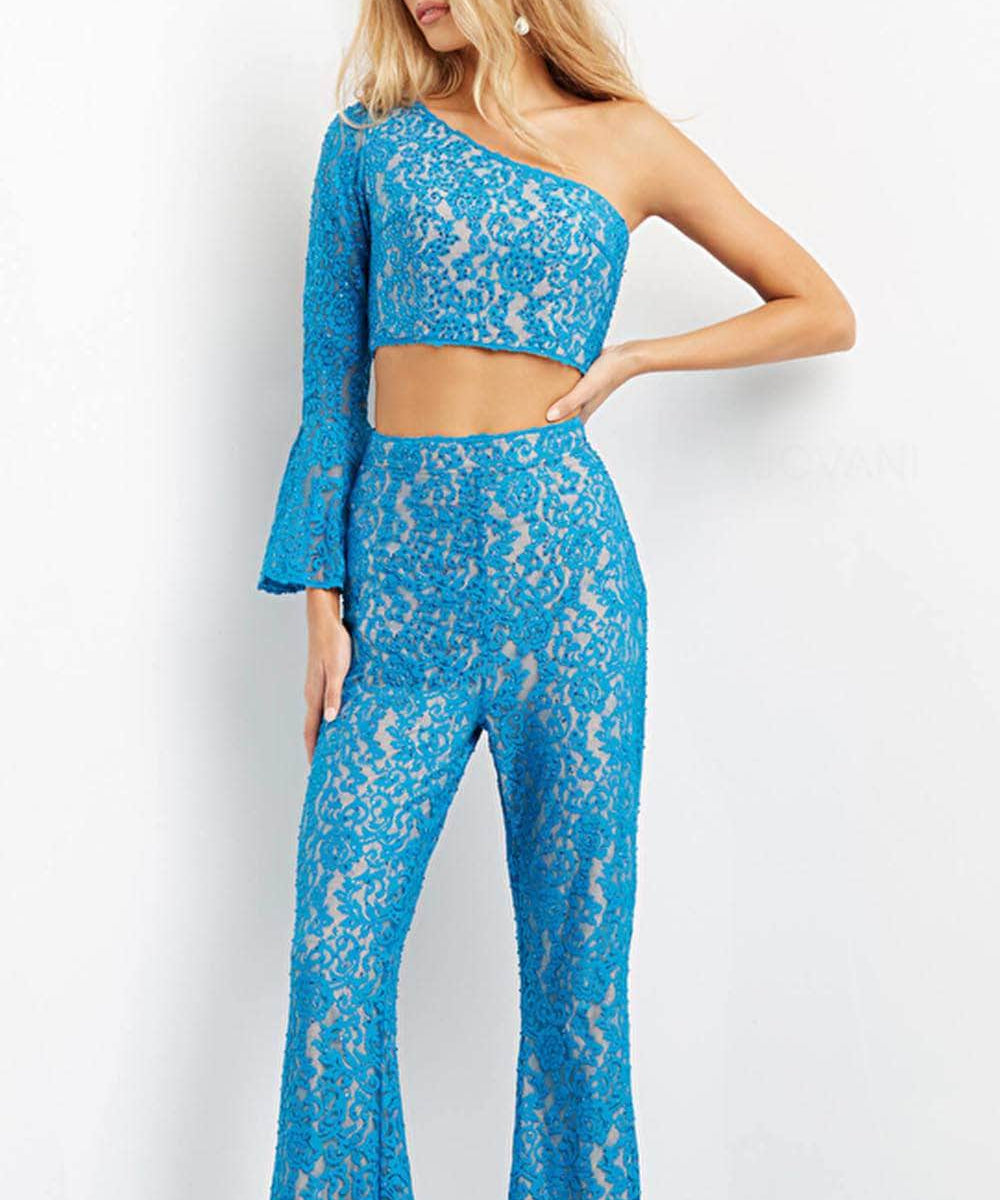 A woman modeling a bright blue lace two-piece outfit consisting of a one-shoulder long-sleeve crop top and matching high-waisted flared pants, photographed against a plain white studio background.