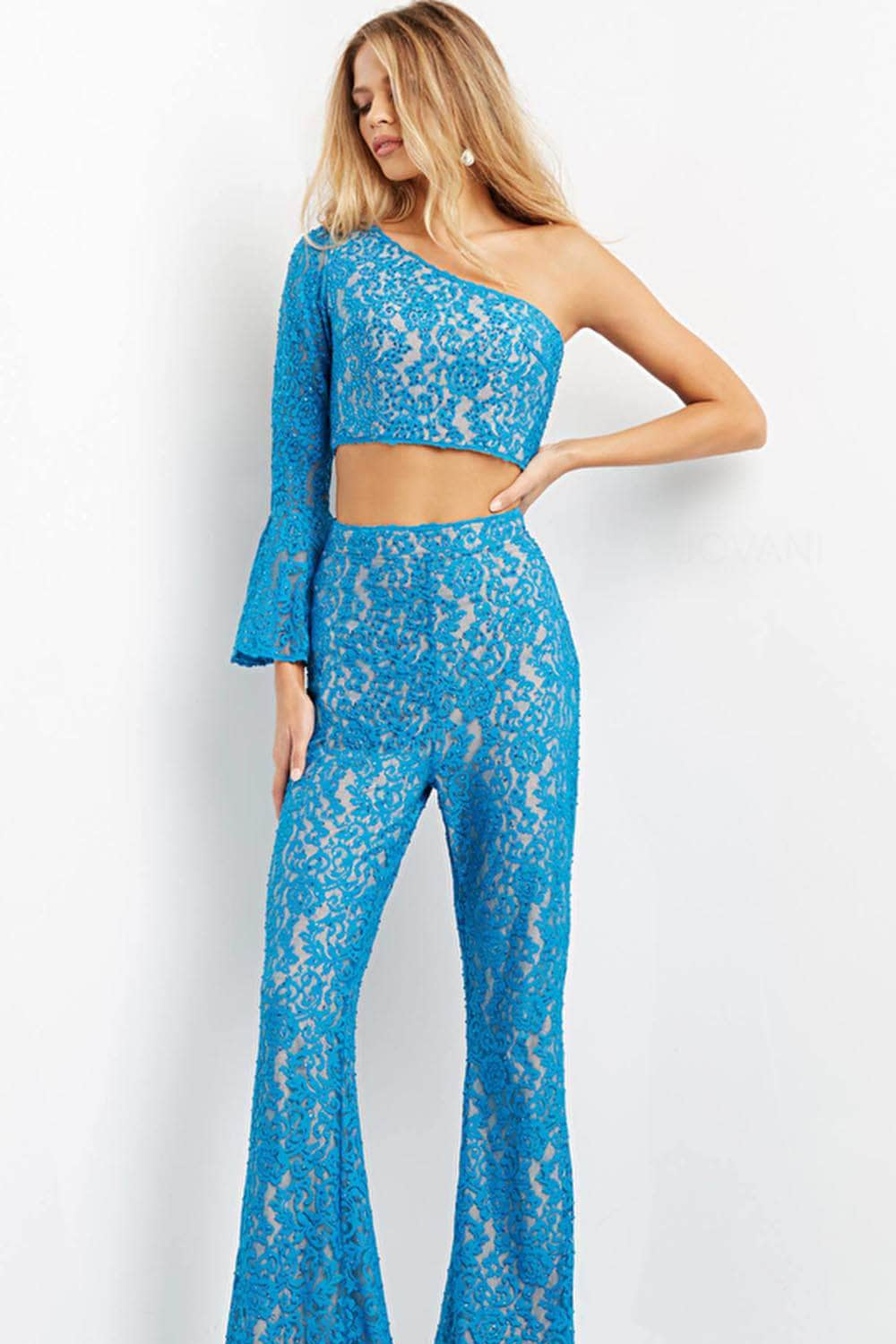 A woman modeling a bright blue lace two-piece outfit consisting of a one-shoulder long-sleeve crop top and matching high-waisted flared pants, photographed against a plain white studio background.