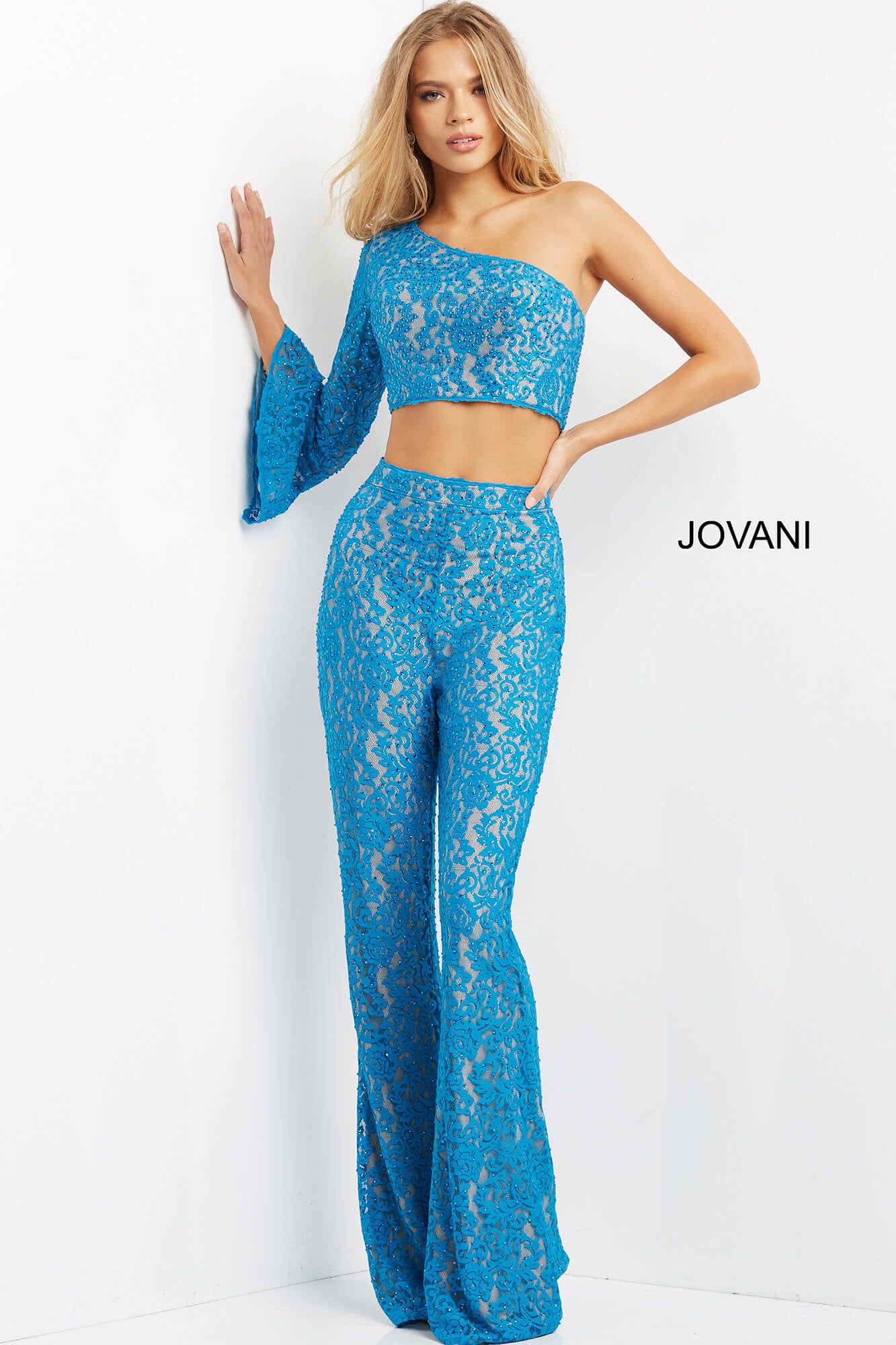 A woman models a bright blue lace two-piece outfit consisting of a one-shoulder crop top with long flared sleeves and matching high-waisted flared pants. The brand name 'JOVANI' is printed on the right side of the image against a plain white studio background.