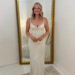 A woman stands in front of a large gold-framed floor mirror wearing a sleeveless, floor-length cream or off-white gown with vertical beadwork and fringe details. The photo is taken in a bright indoor room with white walls and a light rug.