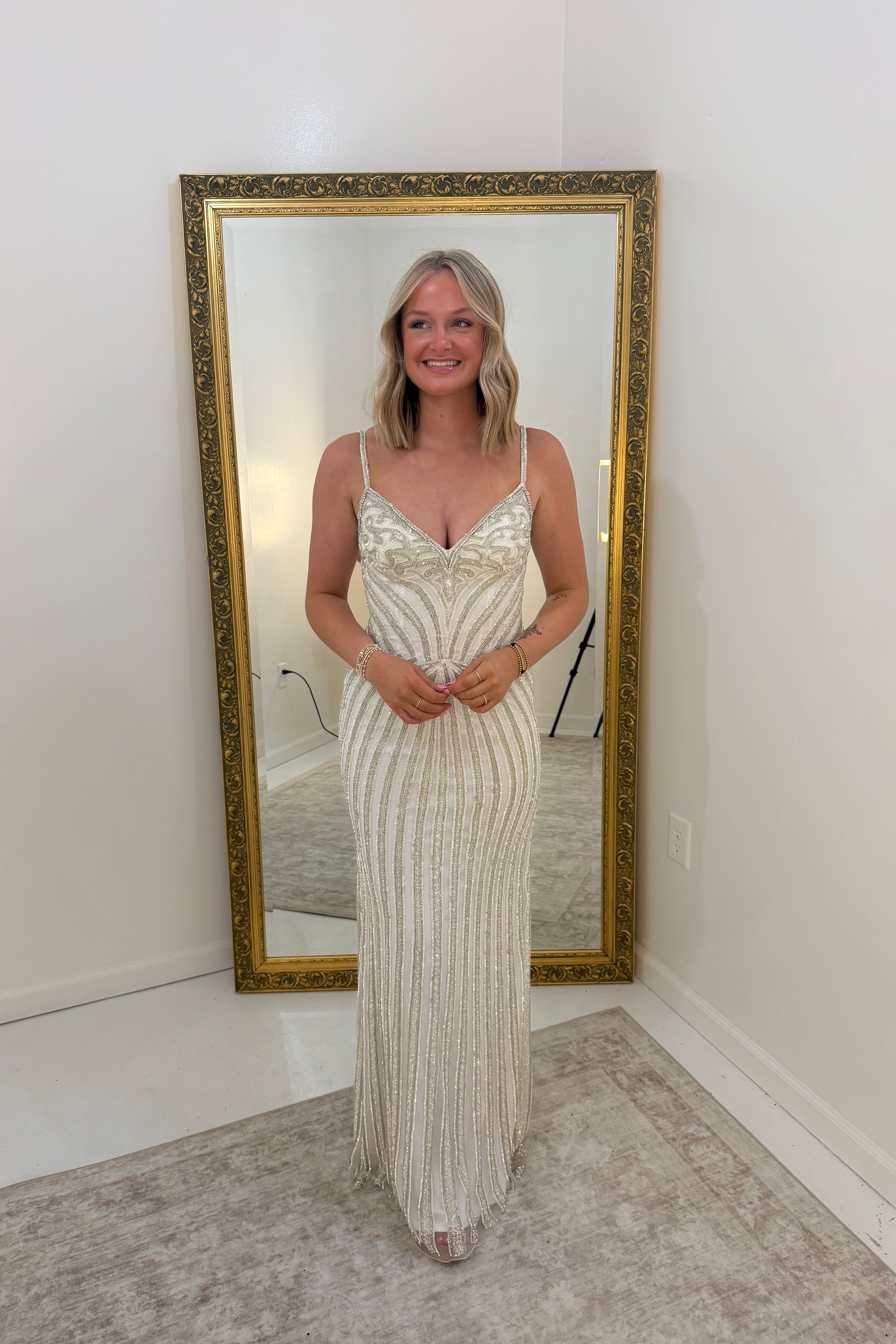 A woman stands in front of a large gold-framed floor mirror wearing a sleeveless, floor-length cream or off-white gown with vertical beadwork and fringe details. The photo is taken in a bright indoor room with white walls and a light rug.