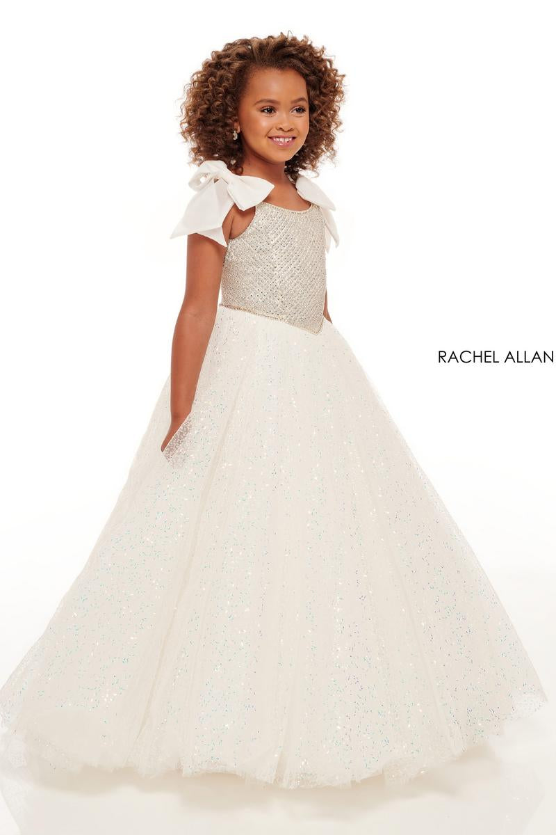 A young girl with curly brown hair wearing a white ball gown with a textured bodice, bow details on the shoulders, and a full tulle skirt, shown against a plain white background with the brand name 'RACHEL ALLAN' printed on the right.