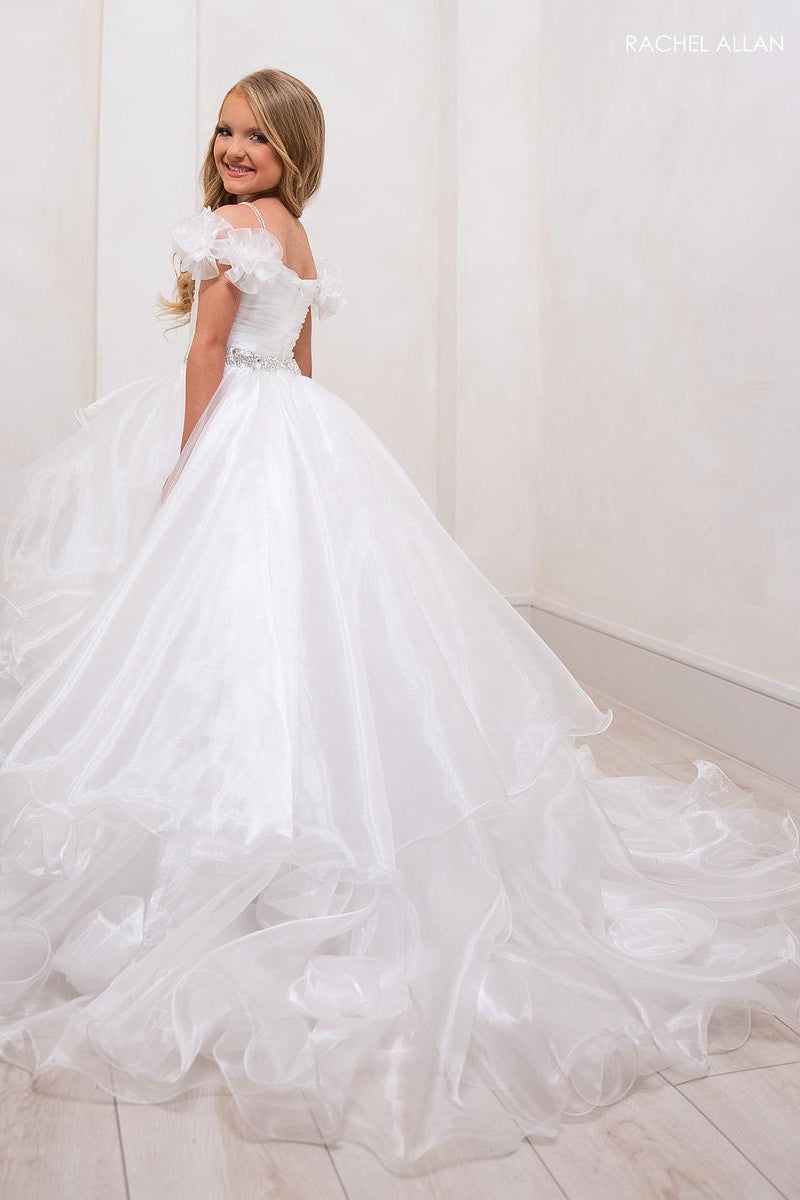 A young girl wearing a white off-the-shoulder ball gown with a full tulle skirt and long train, standing in a bright indoor room with cream walls and light wood flooring. The brand name 'RACHEL ALLAN' appears in the top right corner.