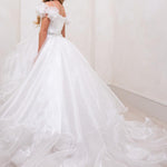 A young girl wearing a white off-the-shoulder ball gown with a full tulle skirt and long train, standing in a bright indoor room with cream walls and light wood flooring. The brand name 'RACHEL ALLAN' appears in the top right corner.