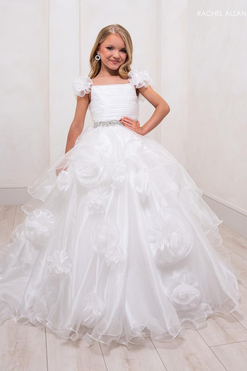 A young girl wearing a white ball gown with 3D floral appliqués and puff sleeves, standing in a bright room with light-colored walls and wood-look flooring. The brand name 'RACHEL ALLAN' is visible in the top right corner.