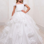 A young girl wearing a white ball gown with 3D floral appliqués and puff sleeves, standing in a bright room with light-colored walls and wood-look flooring. The brand name 'RACHEL ALLAN' is visible in the top right corner.