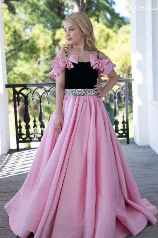 A young blonde girl models a formal ball gown with a black sweetheart bodice, pink off-the-shoulder ruffle sleeves, and a sparkling silver embellished waistband, set against a porch background with green trees behind.