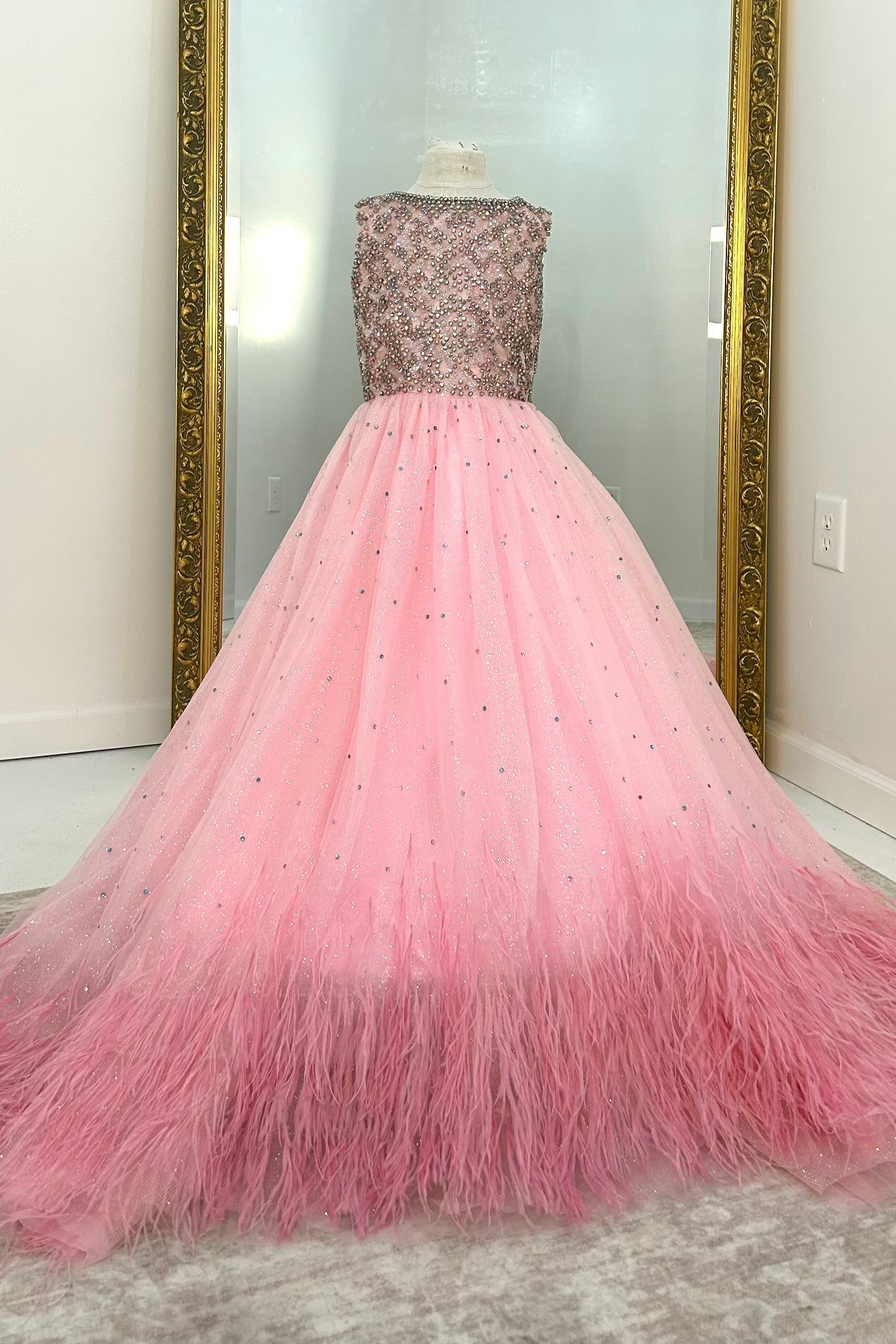 A pink ball gown with a beaded bodice and a voluminous tulle skirt fading into pink feathers at the hem, displayed on a dress form in front of a mirror.