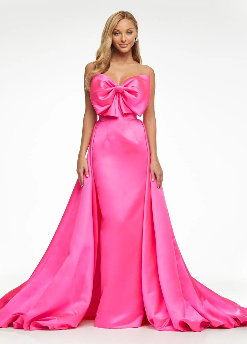 A woman modeling a bright pink strapless evening gown with a large bow at the sweetheart neckline and a full satin skirt with a slight train, photographed against a plain white studio background.