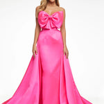 A woman modeling a bright pink strapless evening gown with a large bow at the sweetheart neckline and a full satin skirt with a slight train, photographed against a plain white studio background.