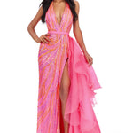 A woman modeling a vibrant pink evening gown with orange and yellow sequined swirls on one side and a flowing sheer pink ruffled overlay on the other, set against a plain white background.