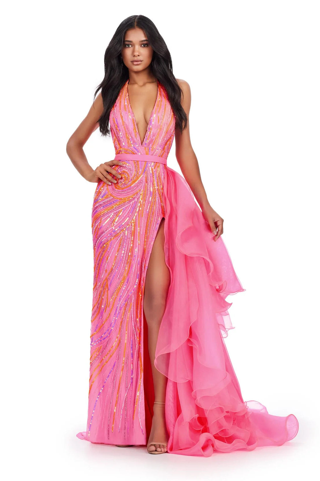 A woman modeling a vibrant pink evening gown with orange and yellow sequined swirls on one side and a flowing sheer pink ruffled overlay on the other, set against a plain white background.