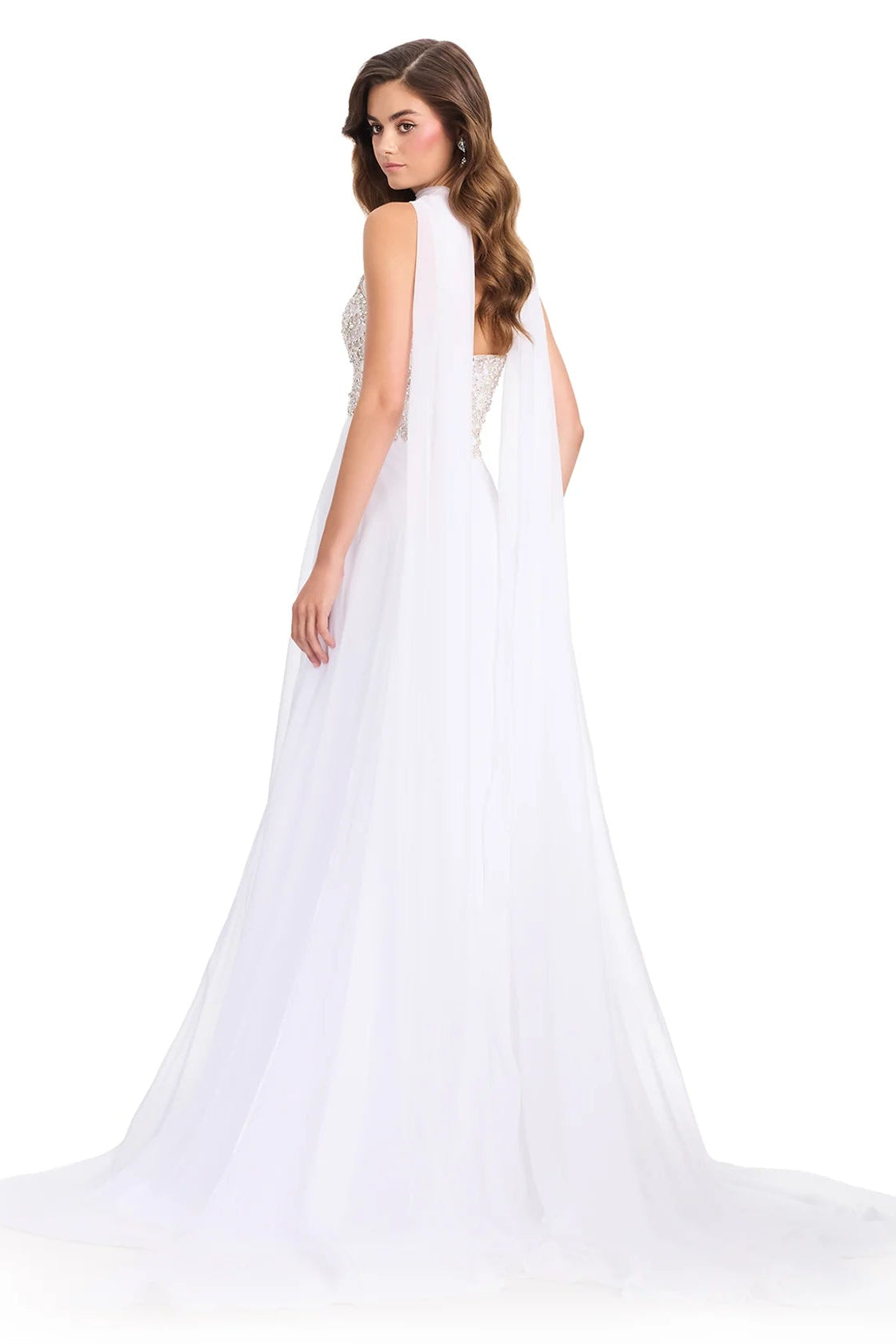 A woman modeling a white bridal gown from the back, featuring a sheer draped cape-style back drape, a beaded open back, and a long flowing tulle train against a plain white studio background.