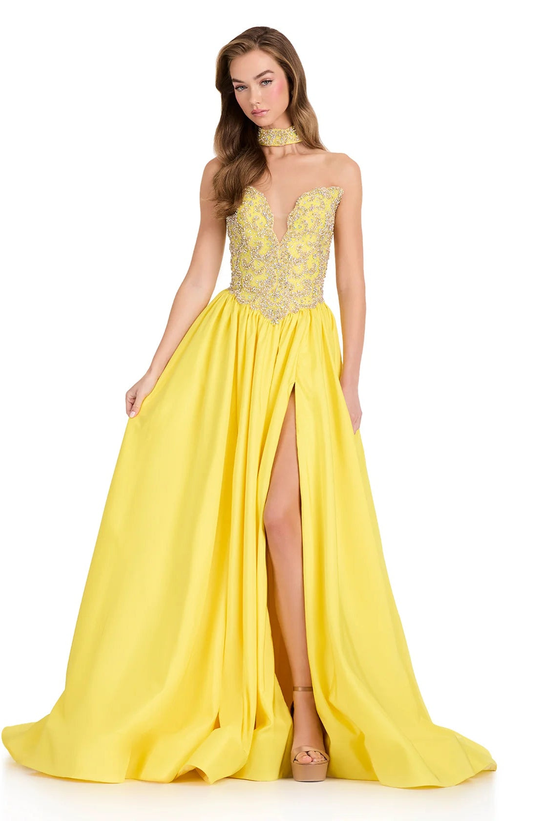 A woman modeling a bright yellow formal gown with a sheer lace bodice, choker neckline, and a thigh slit, photographed against a plain white studio background.