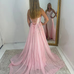 A woman stands with her back to a large gold-framed mirror, wearing a long pink formal gown with a lace-up back and a flowing train. The mirror shows the front of the dress, which appears strapless with a fitted bodice and embellished details.