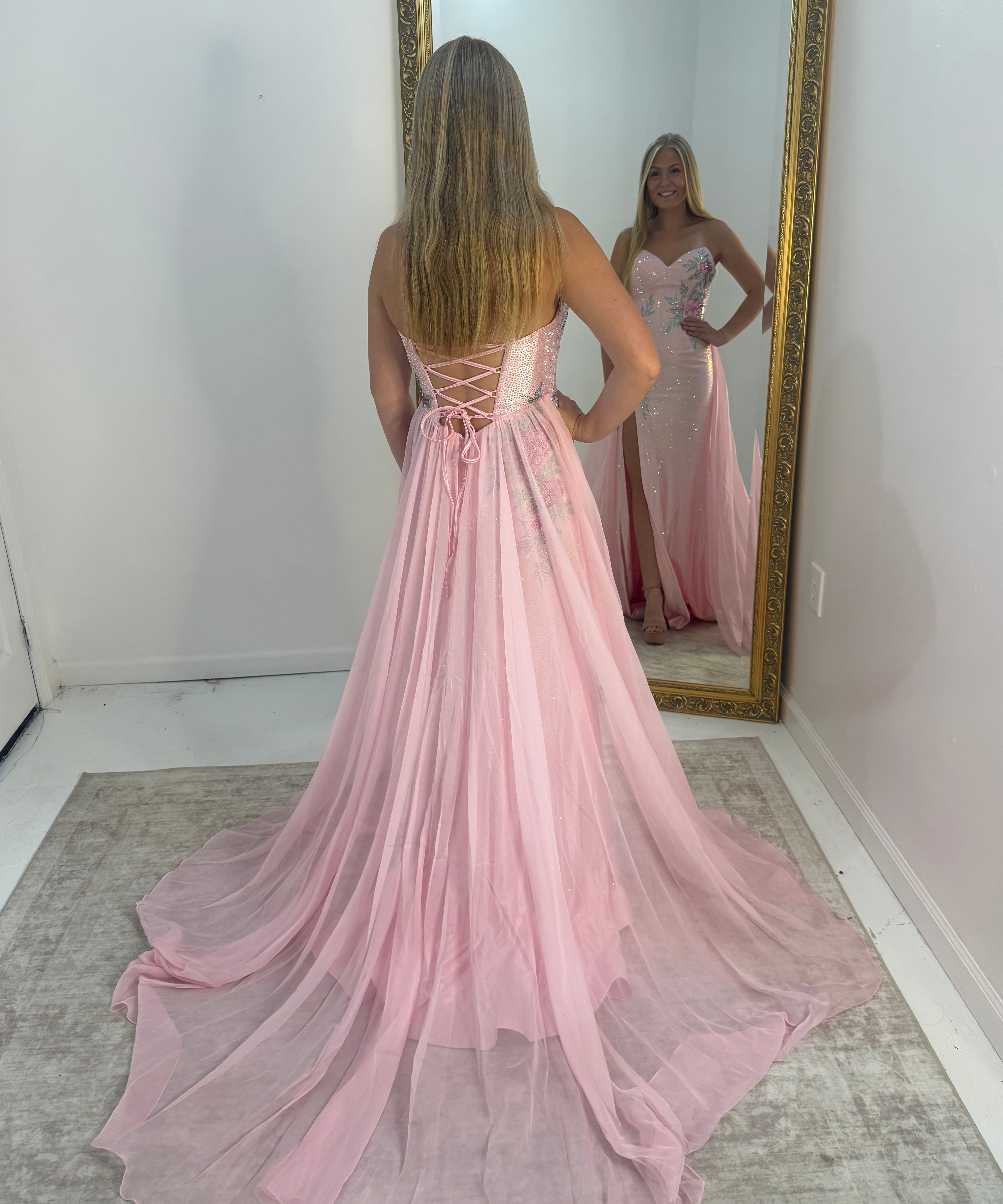 A woman stands with her back to a large gold-framed mirror, wearing a long pink formal gown with a lace-up back and a flowing train. The mirror shows the front of the dress, which appears strapless with a fitted bodice and embellished details.