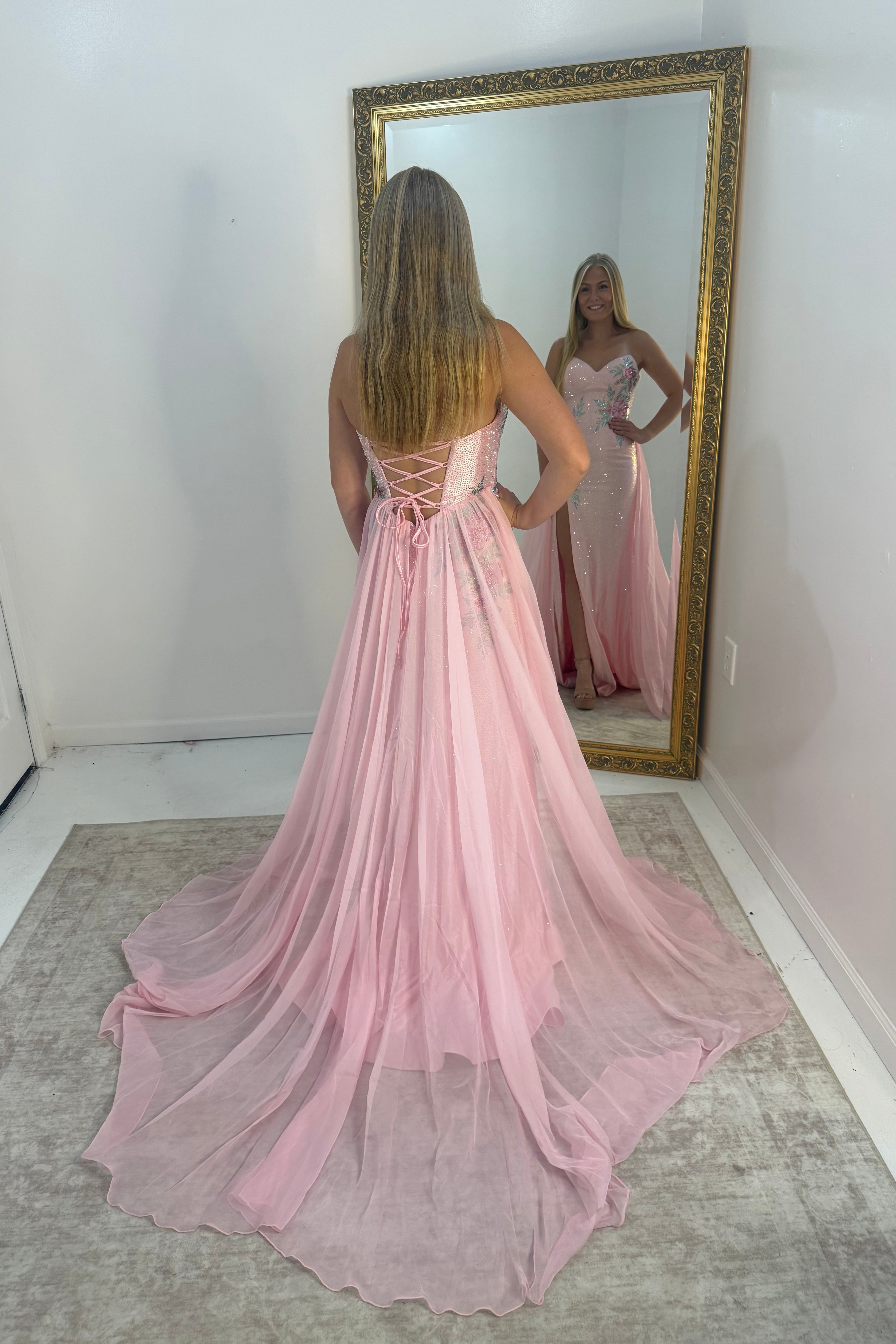 A woman stands with her back to a large gold-framed mirror, wearing a long pink formal gown with a lace-up back and a flowing train. The mirror shows the front of the dress, which appears strapless with a fitted bodice and embellished details.