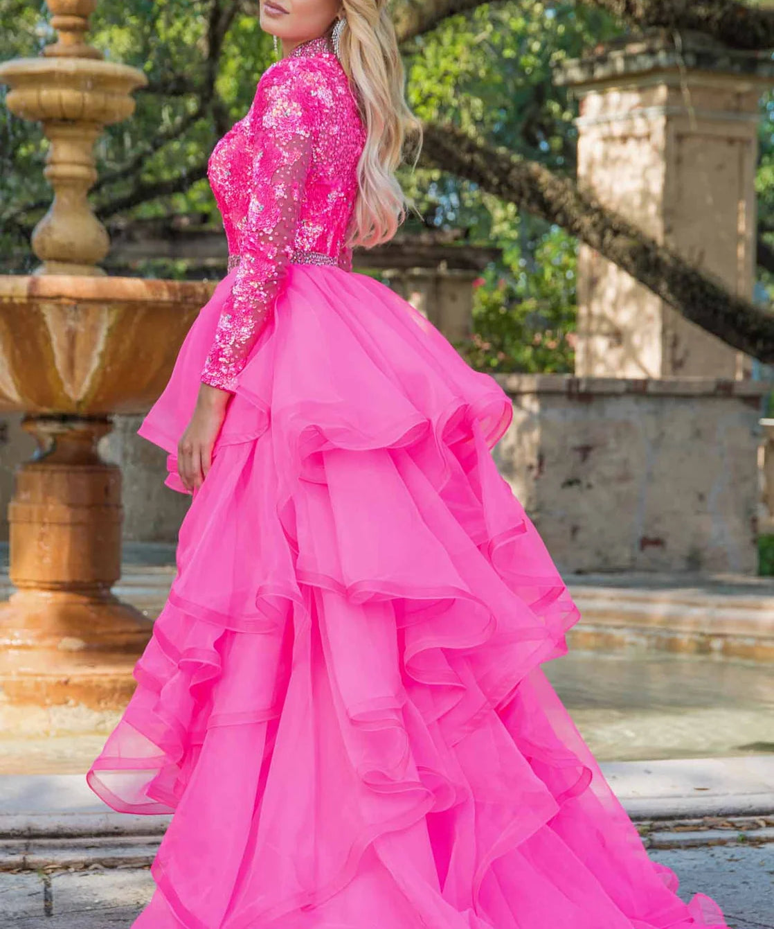 Woman in a pink formal gown standing outdoors with a fountain and trees in the background