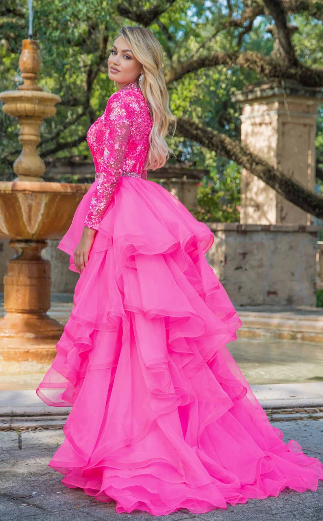 Woman in a pink formal gown standing outdoors with a fountain and trees in the background