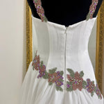 White formal dress displayed on a black mannequin, showing the back view with beaded floral embellishments and embellished shoulder straps.