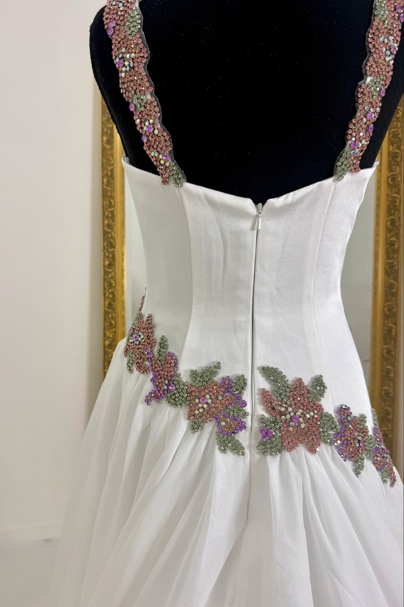 White formal dress displayed on a black mannequin, showing the back view with beaded floral embellishments and embellished shoulder straps.
