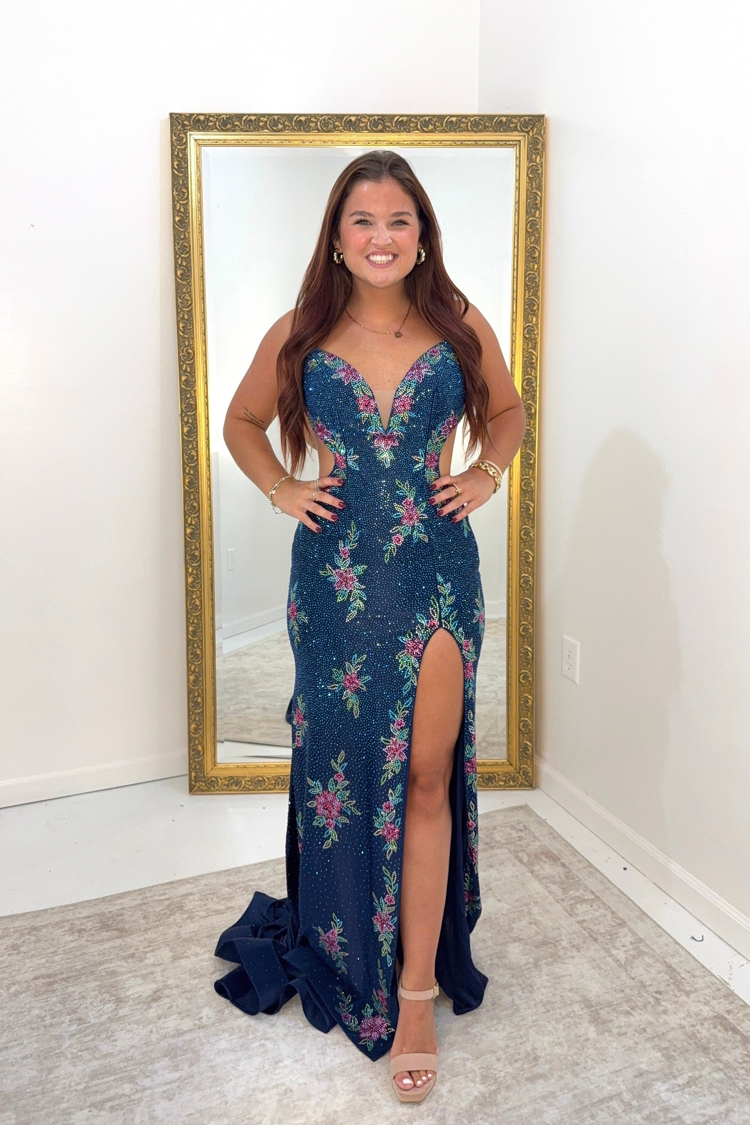 A woman stands indoors wearing a sleeveless navy evening gown with a floral print in pink, green, and blue, featuring a deep V-neckline, fitted silhouette, thigh-high slit, and a small train. She is posed in front of a large gold-framed mirror against a white wall.