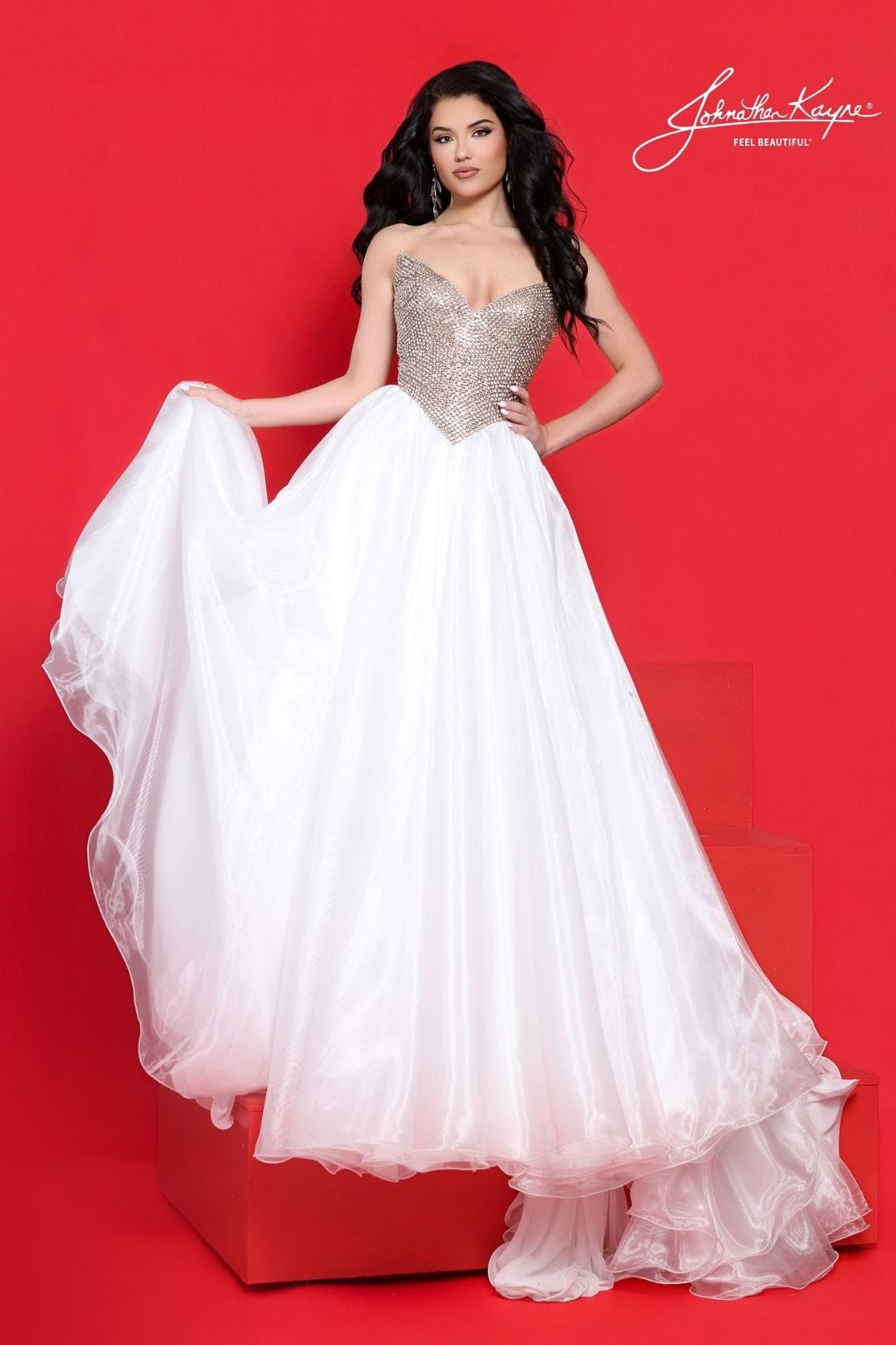 A woman models a strapless ball gown with a sparkling gold-beaded sweetheart bodice and a voluminous white organza skirt, posed against a solid red studio backdrop with a red block base.