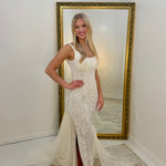 A woman stands indoors wearing an ivory lace mermaid gown with a high slit and long draped train, posed in front of a large gold-framed mirror.