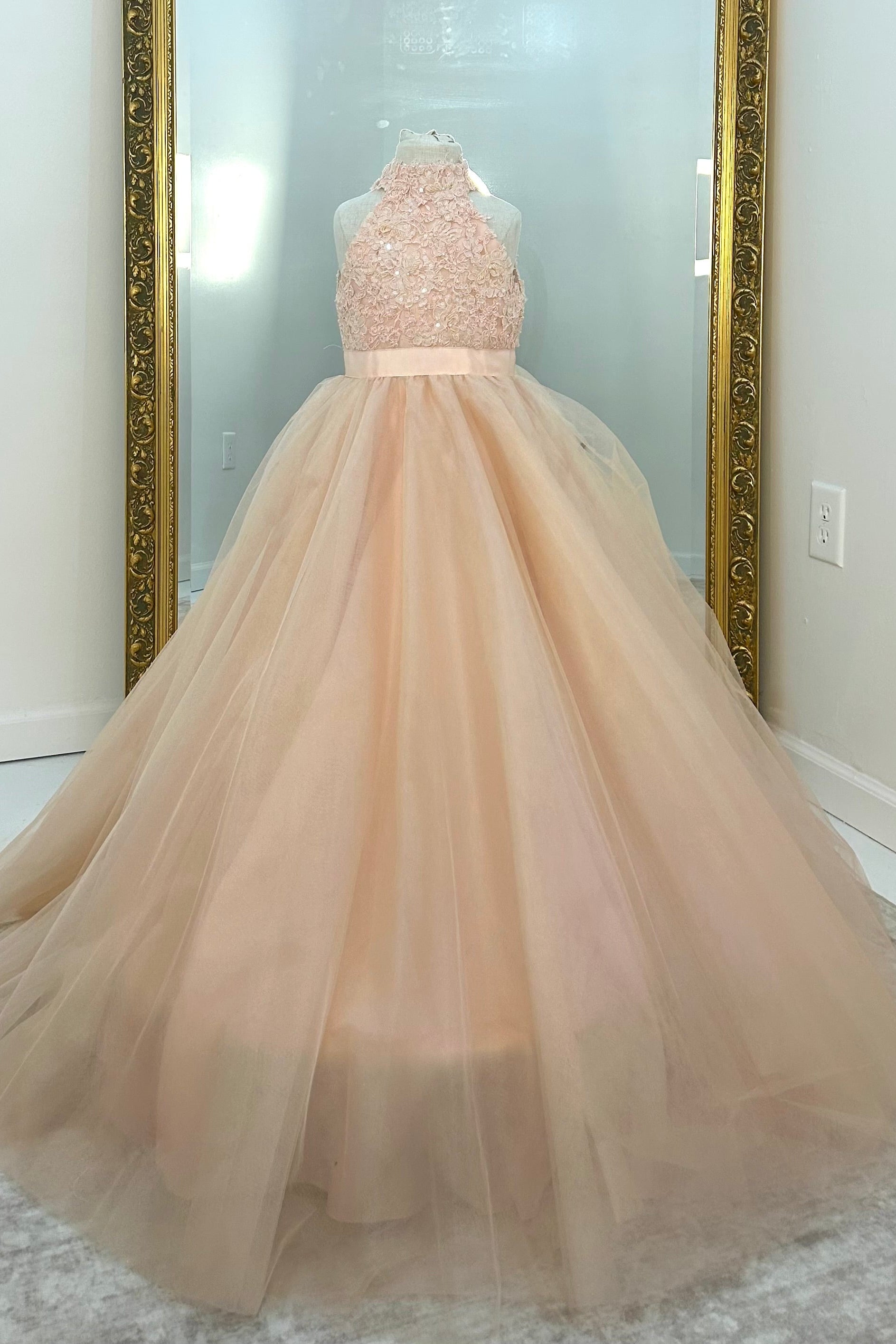 A formal ball gown displayed on a mannequin in front of a large mirror. The dress features a sleeveless, beaded halter-style bodice in a light peach-pink color, connected to a voluminous, layered tulle skirt in a matching pale blush tone.