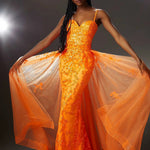 A model wearing a vibrant orange evening gown with sequined floral detailing and a sheer, flowing overskirt, posed against a dark studio backdrop with dramatic lighting.