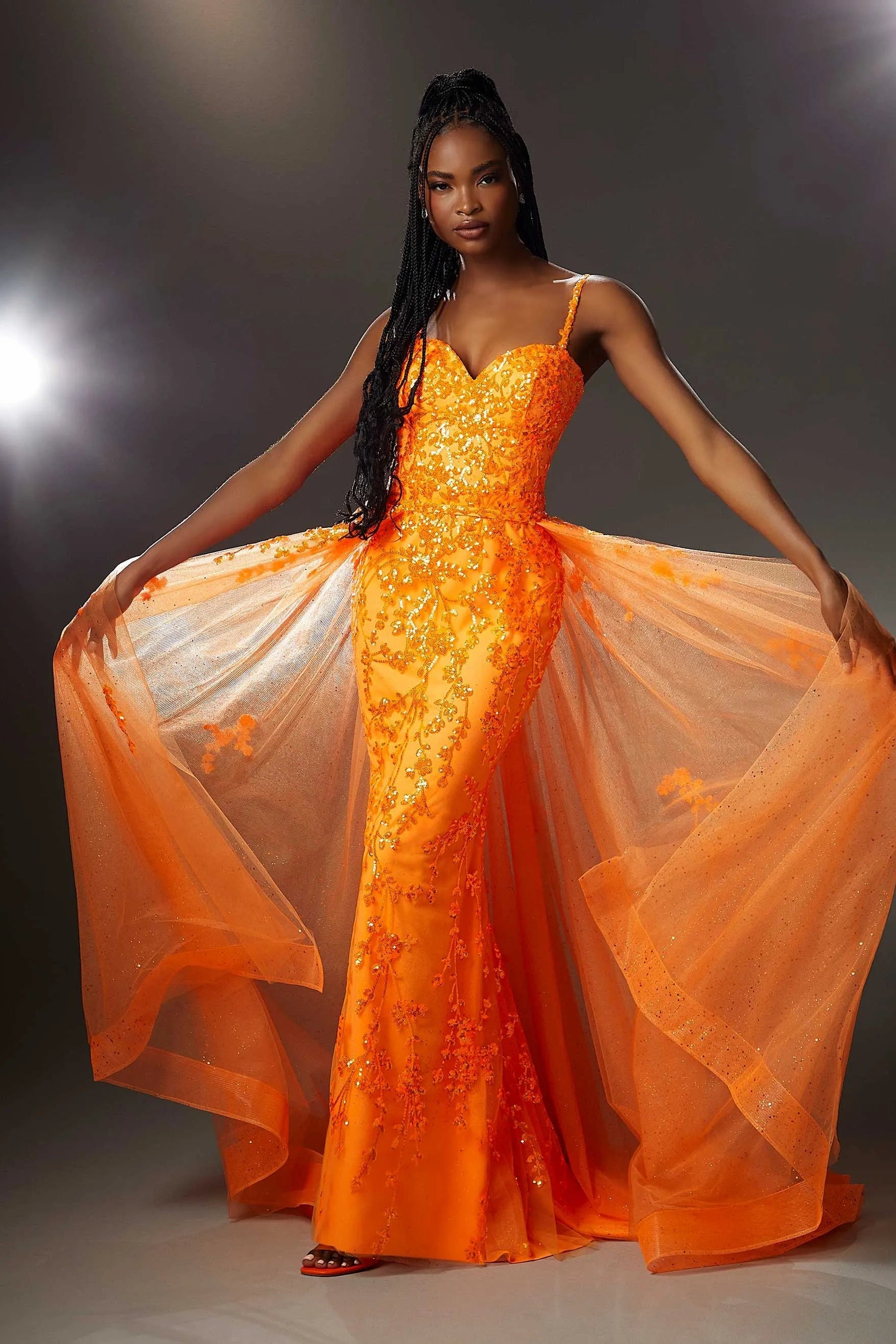 A model wearing a vibrant orange evening gown with sequined floral detailing and a sheer, flowing overskirt, posed against a dark studio backdrop with dramatic lighting.