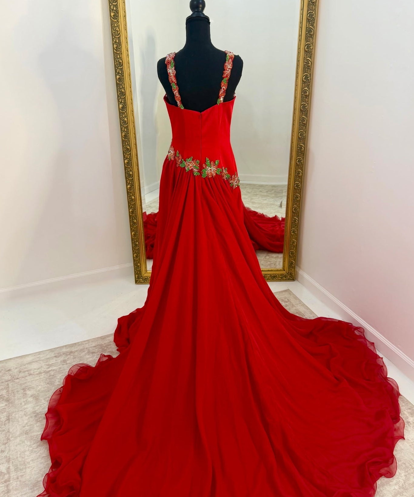 A bright red formal gown displayed on a black mannequin in front of a large gold-framed mirror, showing the back view with a long train on a light-colored floor.