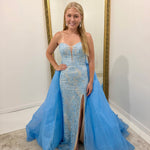A woman stands indoors wearing a strapless light blue evening gown with a fitted lace-embroidered bodice, thigh-high slit, and a voluminous flowing tulle overskirt. She is posed in front of a large gold-framed mirror against a white wall.