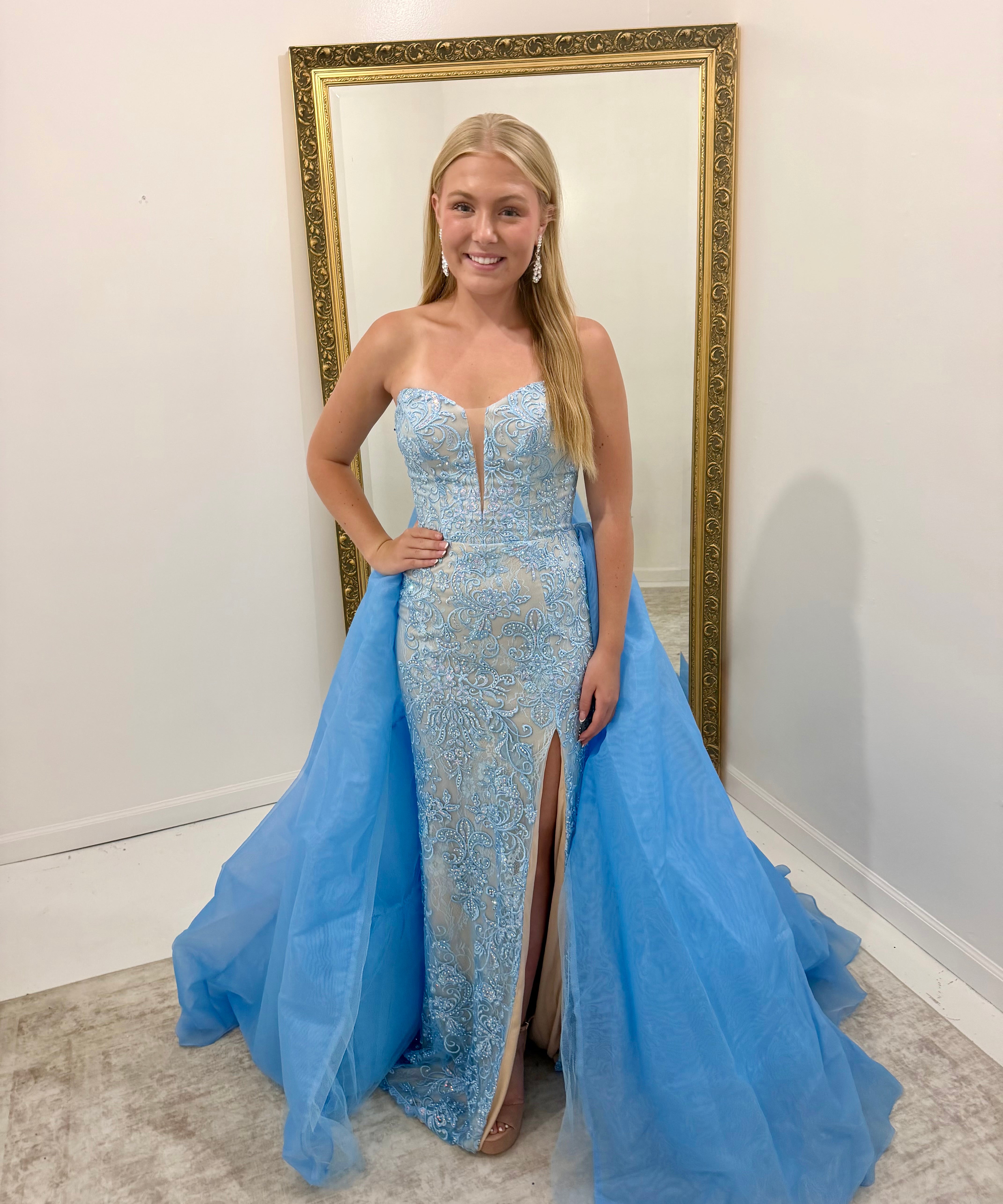 A woman stands indoors wearing a strapless light blue evening gown with a fitted lace-embroidered bodice, thigh-high slit, and a voluminous flowing tulle overskirt. She is posed in front of a large gold-framed mirror against a white wall.