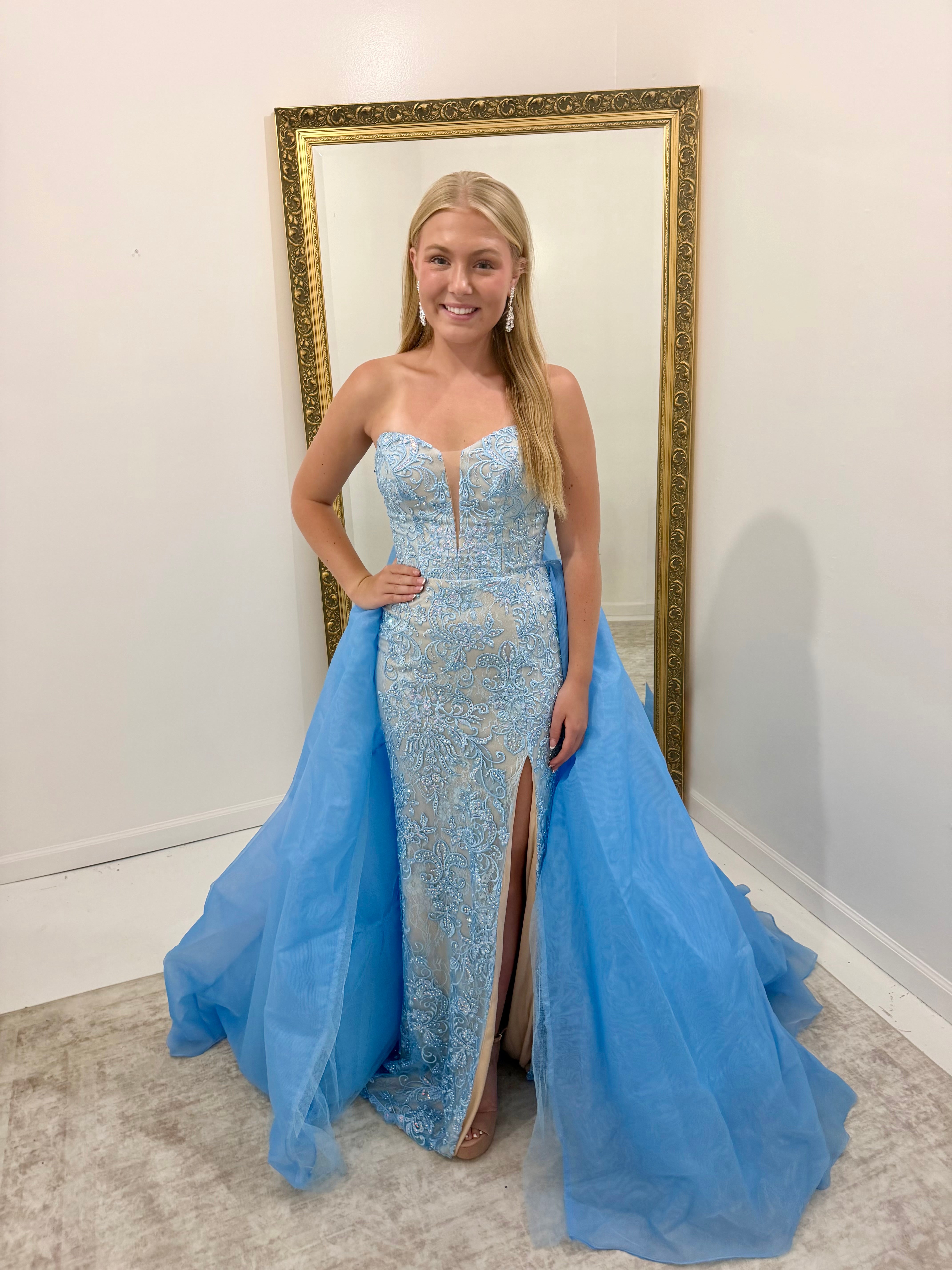 A woman stands indoors wearing a strapless light blue evening gown with a fitted lace-embroidered bodice, thigh-high slit, and a voluminous flowing tulle overskirt. She is posed in front of a large gold-framed mirror against a white wall.