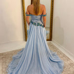 A woman stands with her back to the camera wearing a light blue off-the-shoulder ball gown with a long train, posing in front of a large gold-framed mirror in a room with white walls and a beige rug.