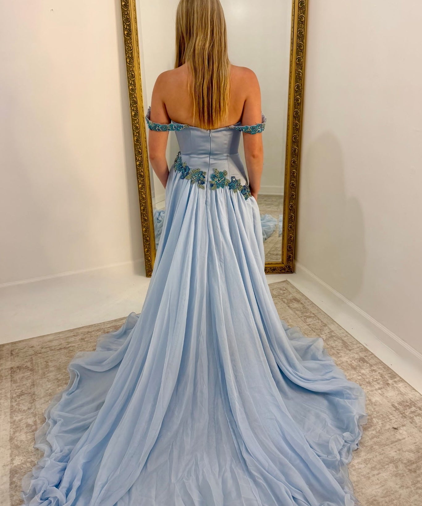 A woman stands with her back to the camera wearing a light blue off-the-shoulder ball gown with a long train, posing in front of a large gold-framed mirror in a room with white walls and a beige rug.