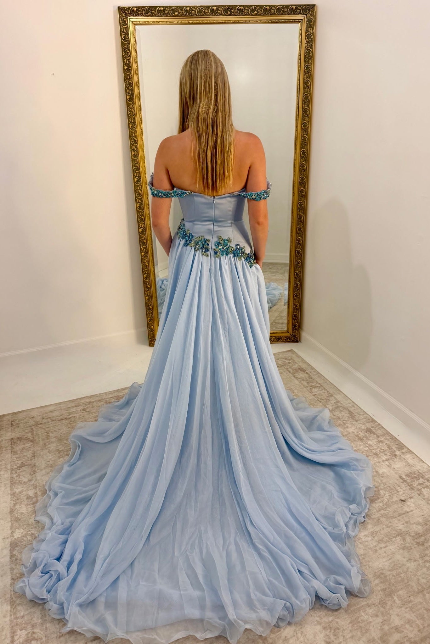 A woman stands with her back to the camera wearing a light blue off-the-shoulder ball gown with a long train, posing in front of a large gold-framed mirror in a room with white walls and a beige rug.