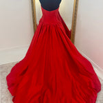 A bright red ball gown displayed on a black dress form mannequin, positioned in front of a large gold-framed mirror on a light beige carpeted floor.