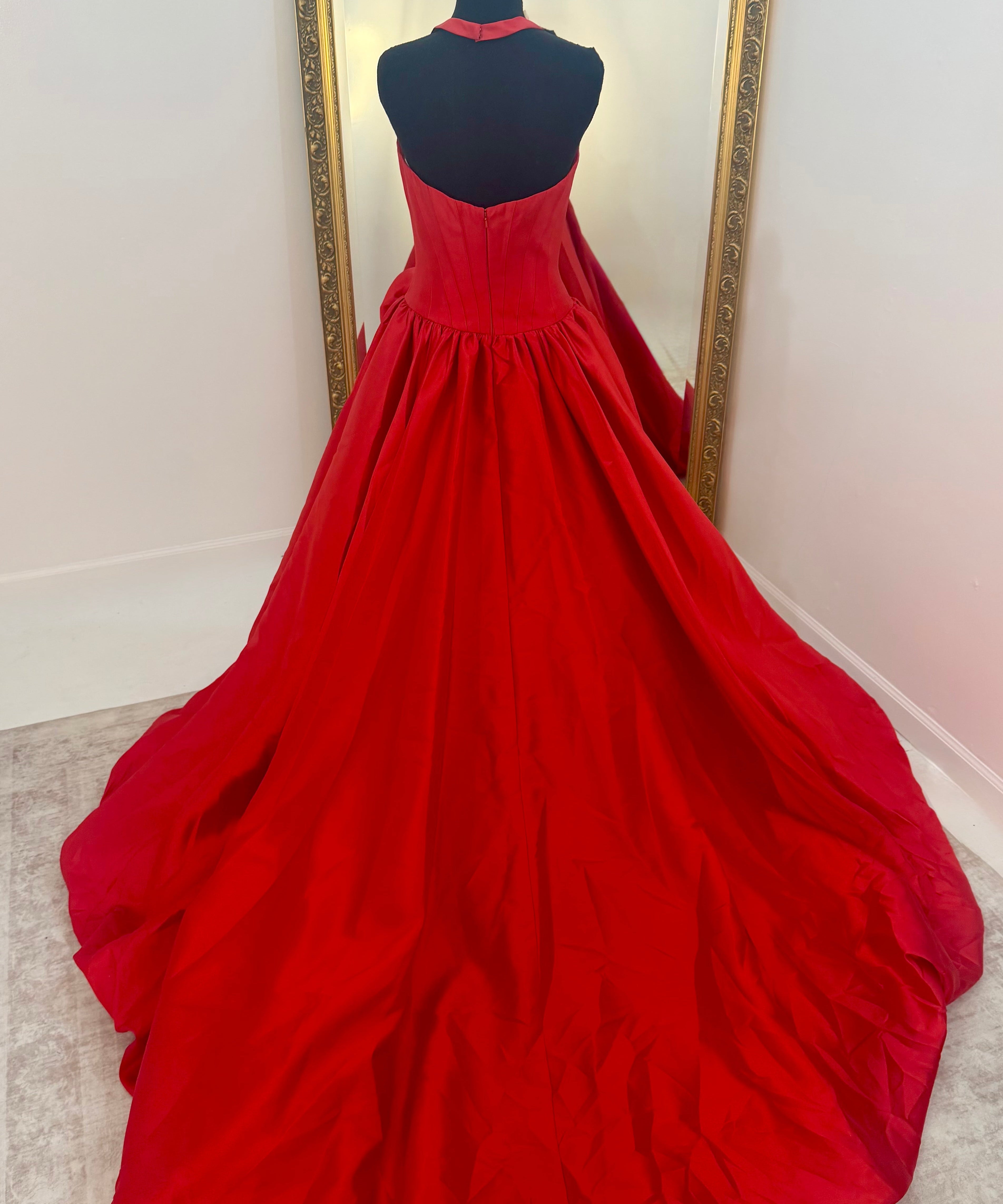 A bright red ball gown displayed on a black dress form mannequin, positioned in front of a large gold-framed mirror on a light beige carpeted floor.