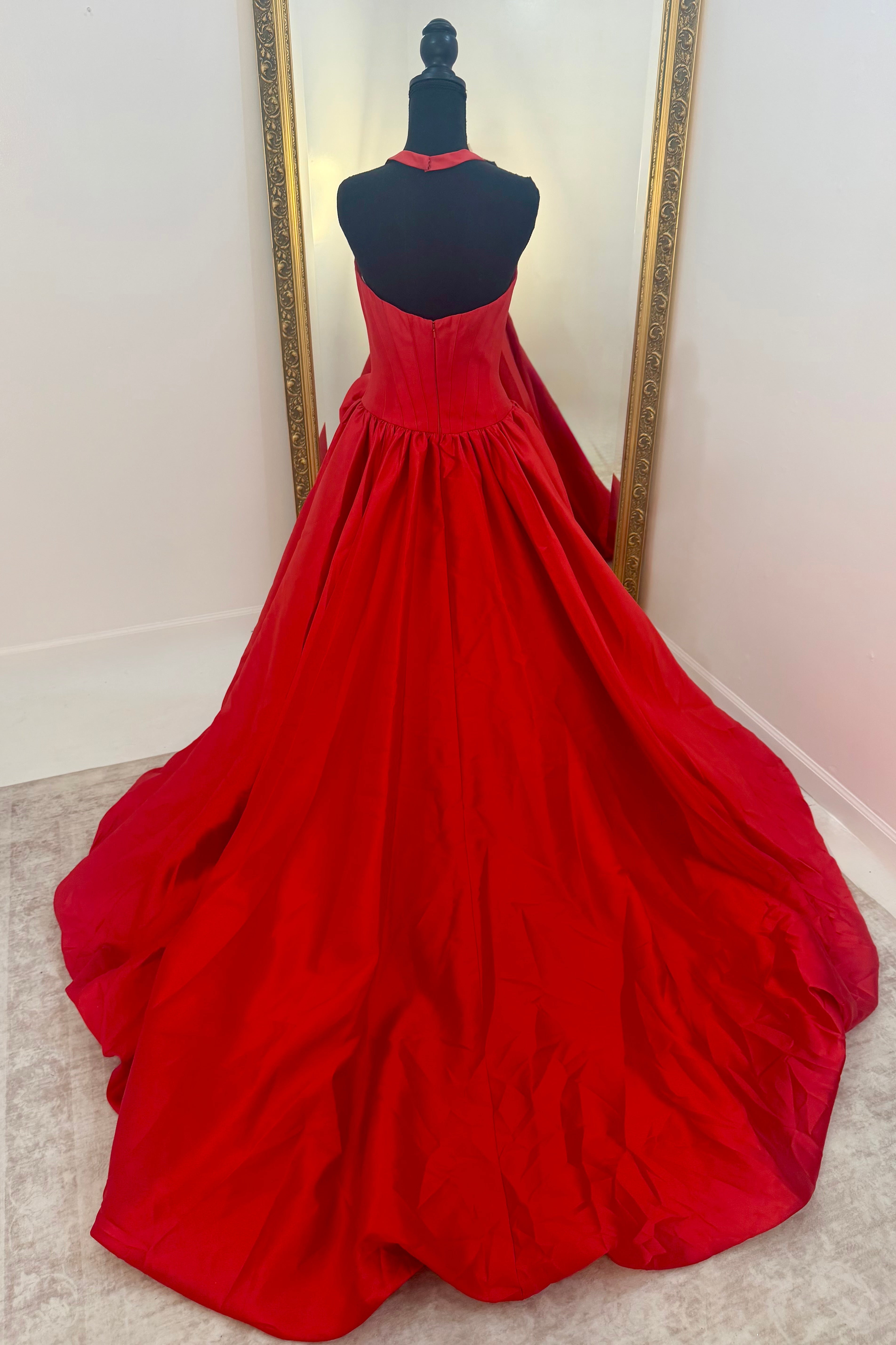 A bright red ball gown displayed on a black dress form mannequin, positioned in front of a large gold-framed mirror on a light beige carpeted floor.