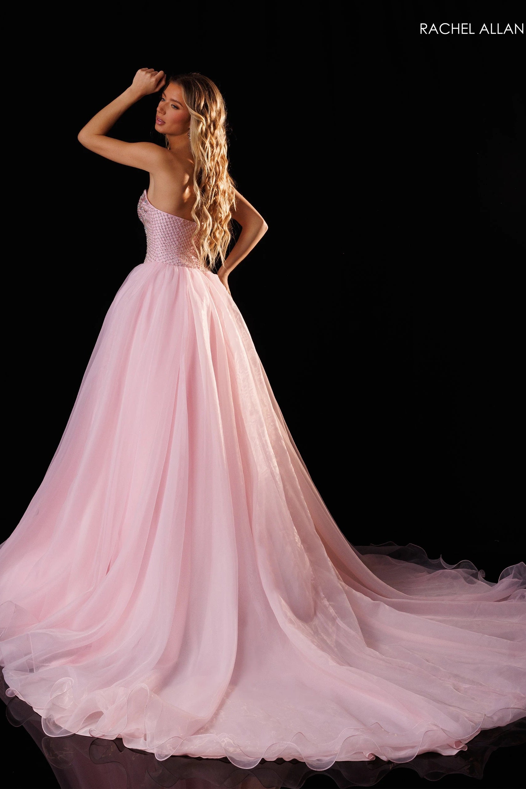 A woman modeling a pale pink strapless ball gown with a full tulle skirt and long train against a black studio background. The brand name 'RACHEL ALLAN' appears in the top right corner.