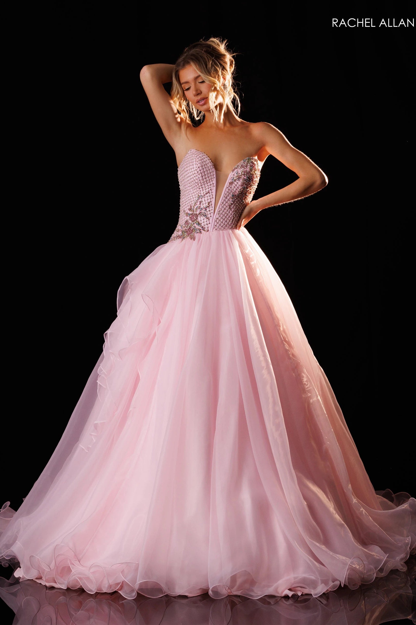 A woman models a pink ball gown with a strapless sweetheart bodice, beaded embellishments, and a voluminous layered tulle skirt against a black studio background. The brand name 'RACHEL ALLAN' is printed in the top right corner.