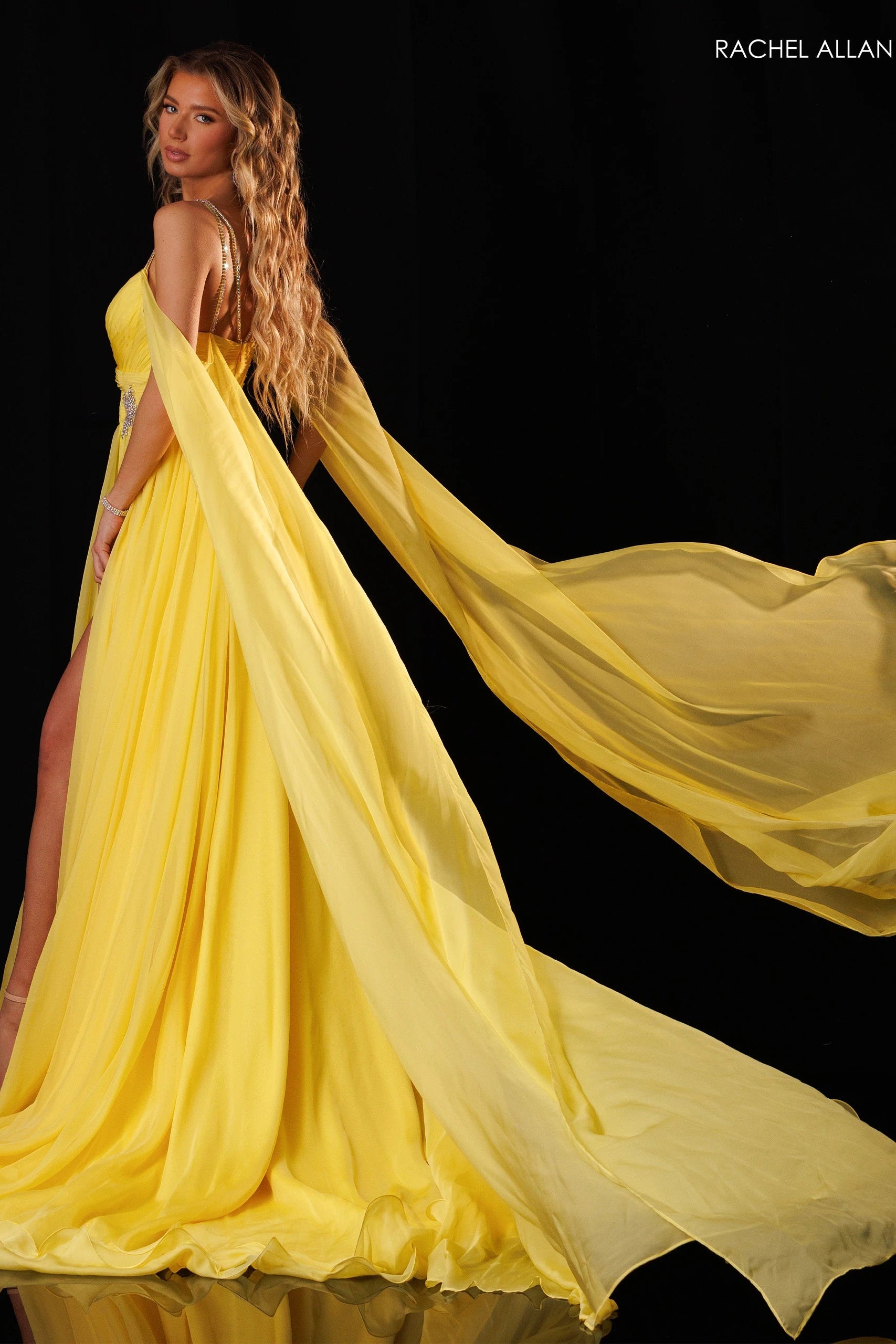A woman modeling a flowing yellow evening gown with a long sheer train against a black background. The brand name 'RACHEL ALLAN' appears in the top right corner.